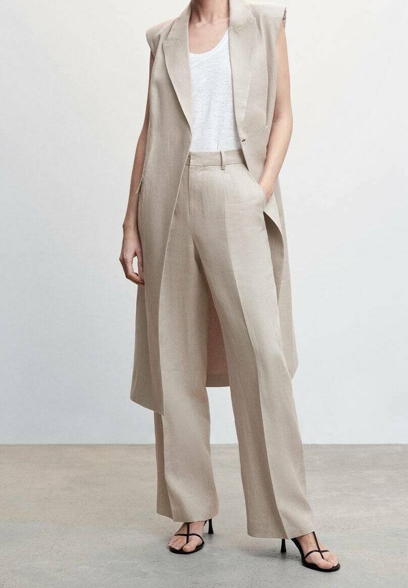 Beżowy garnitur trzyczęściowy z kamizelką bez rękawów, spodniami o szerokich nogawkach i prostą białą koszulką. Sandały mają cienkie czarne paski. Gładka tekstura.