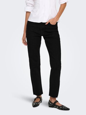 Person trägt schwarze Straight-Leg-Jeans, weißes Hemd mit Knöpfen und hochgekrempelten Ärmeln sowie schwarze mit Nieten besetzte Mary-Jane-Ballerinas und steht mit einer Hand in der Tasche.