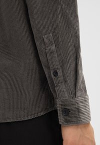 Manica di camicia in corduroy grigio con righe verticali, dotata di bottoni neri e polsino strutturato, che mette in evidenza una consistenza morbida e linee distintive.