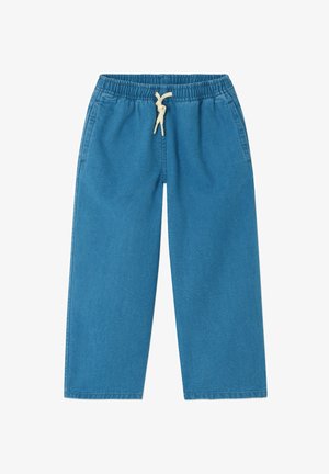 Chlapec stojí a usmieva sa, oblečený v modrej zapínacej bunde, zladených nohaviciach, bielom tričku a béžových sandáloch na svetlom pozadí.