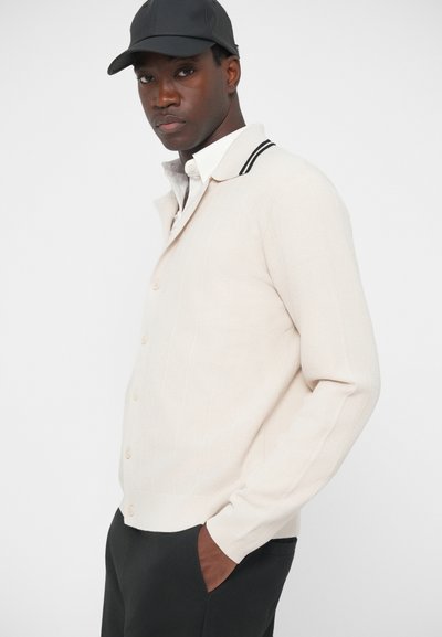 Gaišbeidzīgs adīts kardigāns ar teksturētu rakstu, kuram ir apkakle ar melnām svītrām un pogu aizdare. Uzvilkts virs baltas krekla.