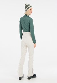 Maglietta verde a maniche lunghe, pantaloni da sci beige con zip laterali e scarponi da sci neri, indossati con un berretto a righe. Design minimalista e forma aderente.
