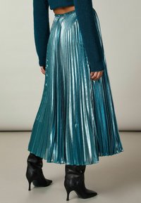 Teal metallisk plisserad maxikjol med hög midja och glänsande struktur, tillsammans med svarta spetsiga klackstövlar.