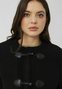 Veste en polaire noire avec des fermetures à bascule et des accents noirs. Texture douce et encolure ronde. Design simple mais fonctionnel.