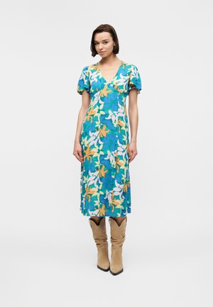 Nainen pukeutuneena sinivihreä-keltavalkoiseen kukkakuosiseen midimekkoon, jossa on liehukkeet hihansuissa, ja ruskeisiin mokkanahkaisiiin polven korkuisiin saappaisiin, seisoo valkoista taustaa vasten.