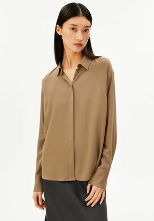 Woman with straight black hair wears loose brown button-up blouse and black skirt, standing against plain white background.