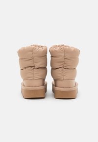 Beige polstrované kotníkové boty s nabraným vrškem, měkkou texturou a plochou, hnědou gumovou podrážkou. Design obsahuje detaily šití.