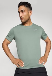 Mees oli salve roheline Nike spordipluus, käed puusadel, vaatab vasakule, lihtsa heleda tausta taustal.