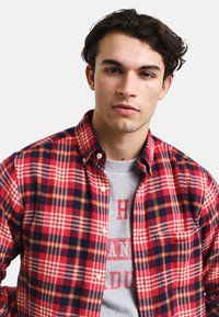 Czerwona i granatowa koszula flanelowa w kratę z kołnierzykiem typu button-down, z białymi akcentami i kieszenią na lewym piersiowym z logo. Nosi się ją na szarej koszulce.