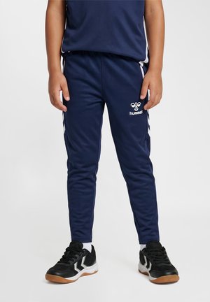 Barn iført marineblå hummel sportbukser med hvide chevronstriber og sorte sneakers med hvide såler, der står på en ensfarvet baggrund.
