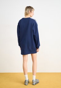 Marinblå oversized sweatshirt i mjukt tyg, med vita ränder längs axlarna, matchad med silverfärgade sportskor och vita strumpor.