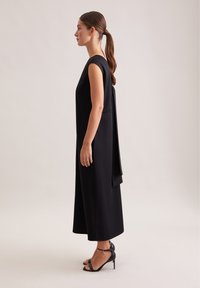 Robe noire sans manches avec un panneau arrière fluide, présentant une silhouette droite et une texture lisse. Associée à des sandales à talons hauts noires.