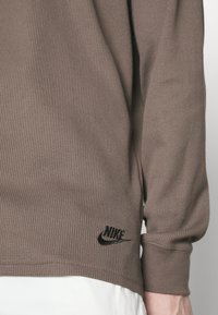 Hnědé žebrované tričko s dlouhým rukávem, kulatým výstřihem a černým logem Nike na spodní levé straně. Materiál má strukturovaný vzhled.