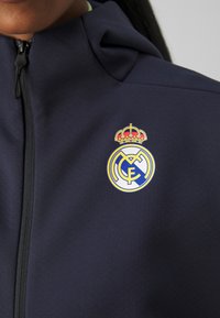 Mörkblå zip-jacka med en rund Real Madrid-logotyp med en krona, i gult, vitt och blått. Mjuk tyg med en slät textur.