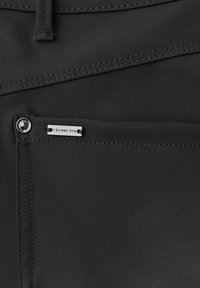 Zbliżenie na czarną tkaninową kieszeń z metalowym guzikiem i małą srebrną metką z napisem "Street One" przyszytą na krawędzi kieszeni.