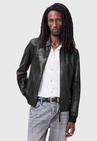 Svart läderbombarjacka med dragkedja framtill, ribbade ärmslut och krage. Bärs över en vit skjorta med knappar och grå jeans.