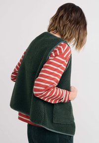 Žalias fliso liemenė su tekstūruota paviršiumi, apvaliu kraštu ir priekiu kišene, dėvima ant raudonos ir baltos juostuotos ilgomis rankovėmis marškinėlių.