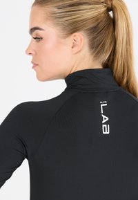 Zwarte sporttop met hoge kraag, gemaakt van een gestructureerde stof en met een wit logo op de achterkant. De zijkant toont een aangesloten ontwerp en lange mouwen.