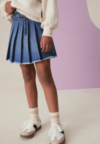 Denim plissérok met franjes, blauwe kleur. G gedragen met witte sneakers en gestreepte sokken. Achtergrond in zachte, gedempte tinten.