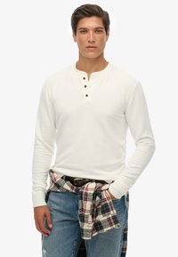 Chemise blanche à manches longues avec un col henley et trois boutons marron, portée avec un jean bleu et une chemise à carreaux nouée à la taille.