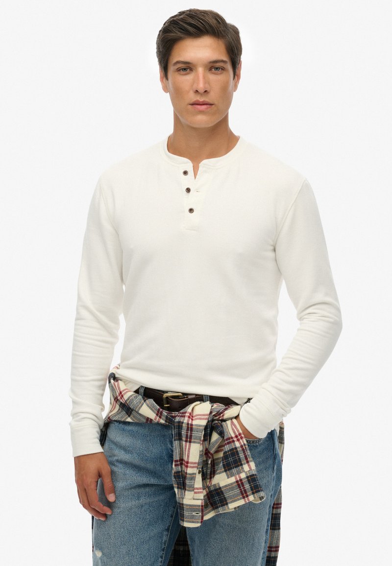 Chemise blanche à manches longues avec un col henley et trois boutons marron, portée avec un jean bleu et une chemise à carreaux nouée à la taille.