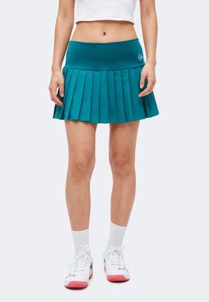 Femme portant une jupe plissée bleu sarcelle, un crop top blanc, des chaussettes blanches et des baskets blanches avec des semelles roses, debout devant un fond uni.