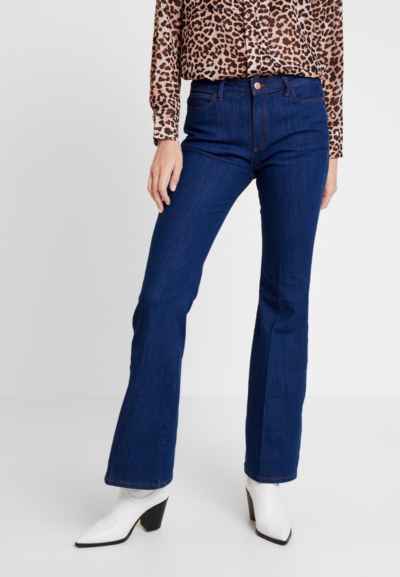 Femme portant un jean bootcut bleu foncé, un chemisier à manches longues imprimé léopard et des bottines blanches à talons, debout devant un fond uni.