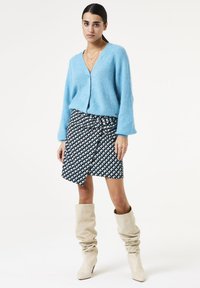 Lichtblauwe gebreide cardigan met een V-hals, gecombineerd met een zwart-wit gemusterde wikkelrok en beige slouch laarzen tot de knie.