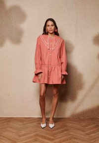 Φόρεμα από βαμβάκι σε ροδακινί χρώμα με μεγάλο λευκό γιακά, κουμπιά στην μπροστινή όψη και μανίκια με βολάν. Φοριέται με λευκά παπούτσια που έχουν μαύρες λεπτομέρειες.