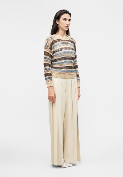 Γυναίκα που στέκεται, φοράει ένα ριγέ πλεκτό πουλόβερ σε μπεζ, μπλε και καφέ αποχρώσεις, φαρδιά εκρού παντελόνια και λευκά μυτερά παπούτσια, μπροστά από ένα μονόχρωμο φόντο.