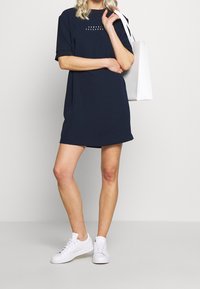 Marinhäftad t-shirtklänning med korta ärmar och rak skärning, med en liten grafisk detalj på framsidan. Matchad med vita sneakers.