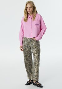 Roze cropped shirt met een borstzak, voorzien van geborduurde tekst. Gecombineerd met beige broek, bedrukt met een zwart luipaardpatroon. Zwarte schoenen.