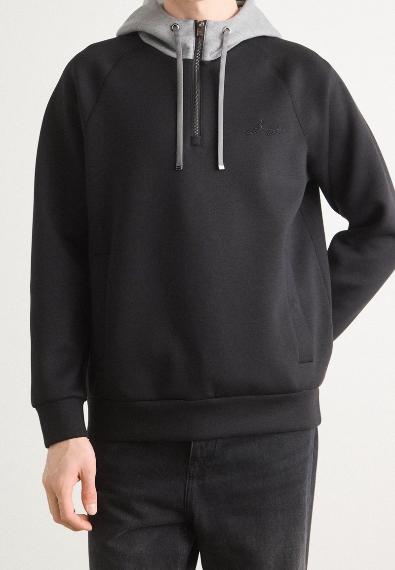 Sudadera negra con capucha gris, con cremallera frontal y cordones. Presenta mangas raglán y un sutil logo en el pecho. Textura suave.