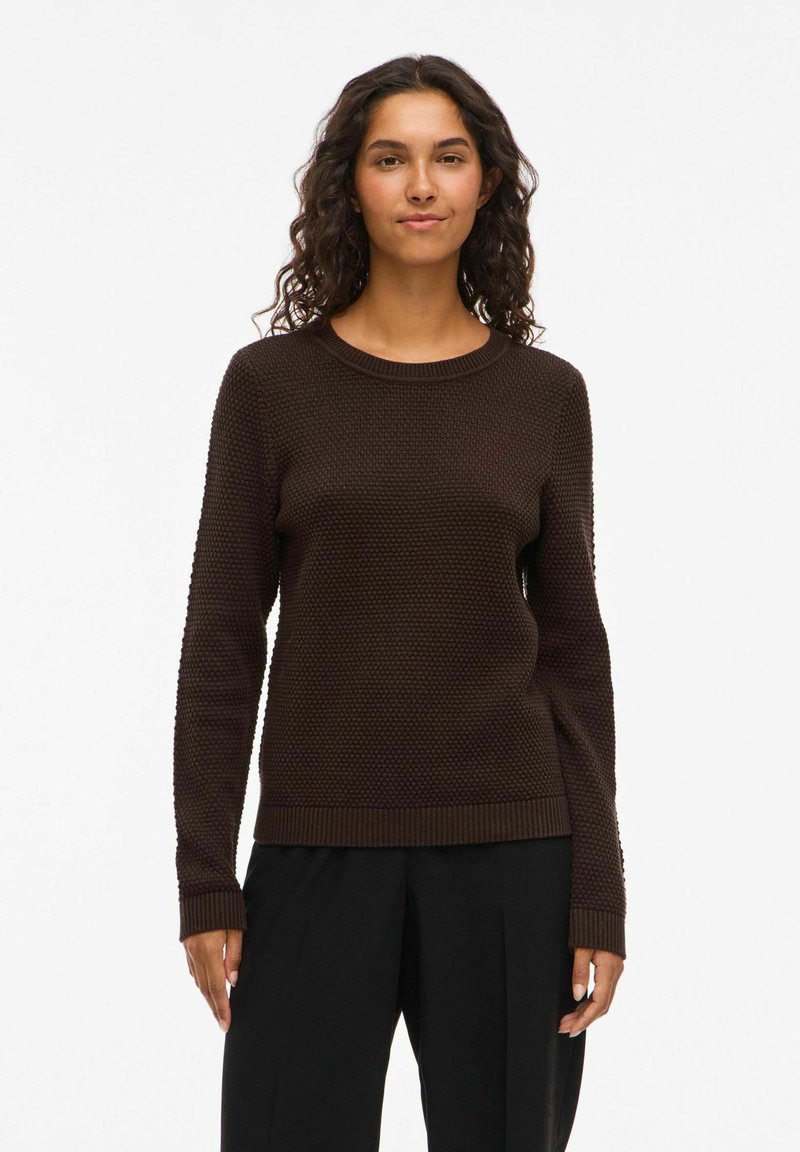 Brūns teksturēts adīts džemperis ar apaļu kakla izgriezumu un garām piedurknēm, ar ribinātām aprocēm un apakšmalu. Valkāts ar melnām biksēm.