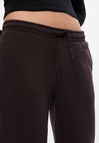 Brūnas sporta bikses, kas izgatavotas no mīksta auduma, ar elastīgu jostasvietu, stingumu un gludu tekstūru, apgriezta skata, kas parāda jostasvietas detalizāciju.