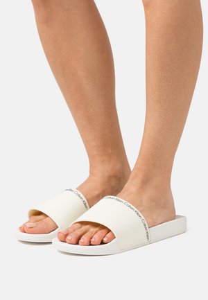 Stopy w białych klapkach Calvin Klein z szerokimi paskami i czarnym logo na jasnym, jednolitym tle.