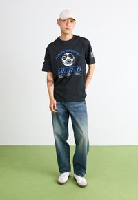 Sort bomuld t-shirt med et fodboldgrafik, blå og hvid tekst, korte ærmer, parret med lyseblå denimjeans og hvide sneakers.