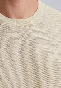 Crème kleurige gebreide trui met een gestructureerd patroon, een ribboord aan de halslijn en een klein geborduurd logo aan de linkerkant.