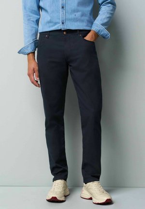 Chino slims bleus marine en coton avec poches avant et détails en bouton marron ; associés à des chaussures de sport crème.