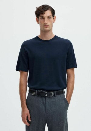 Mann mit kurzen lockigen Haaren, der ein dunkelblaues Kurzarmhemd, graue Hose und einen schwarzen Gürtel trägt, steht vor einem schlichten hellen Hintergrund.