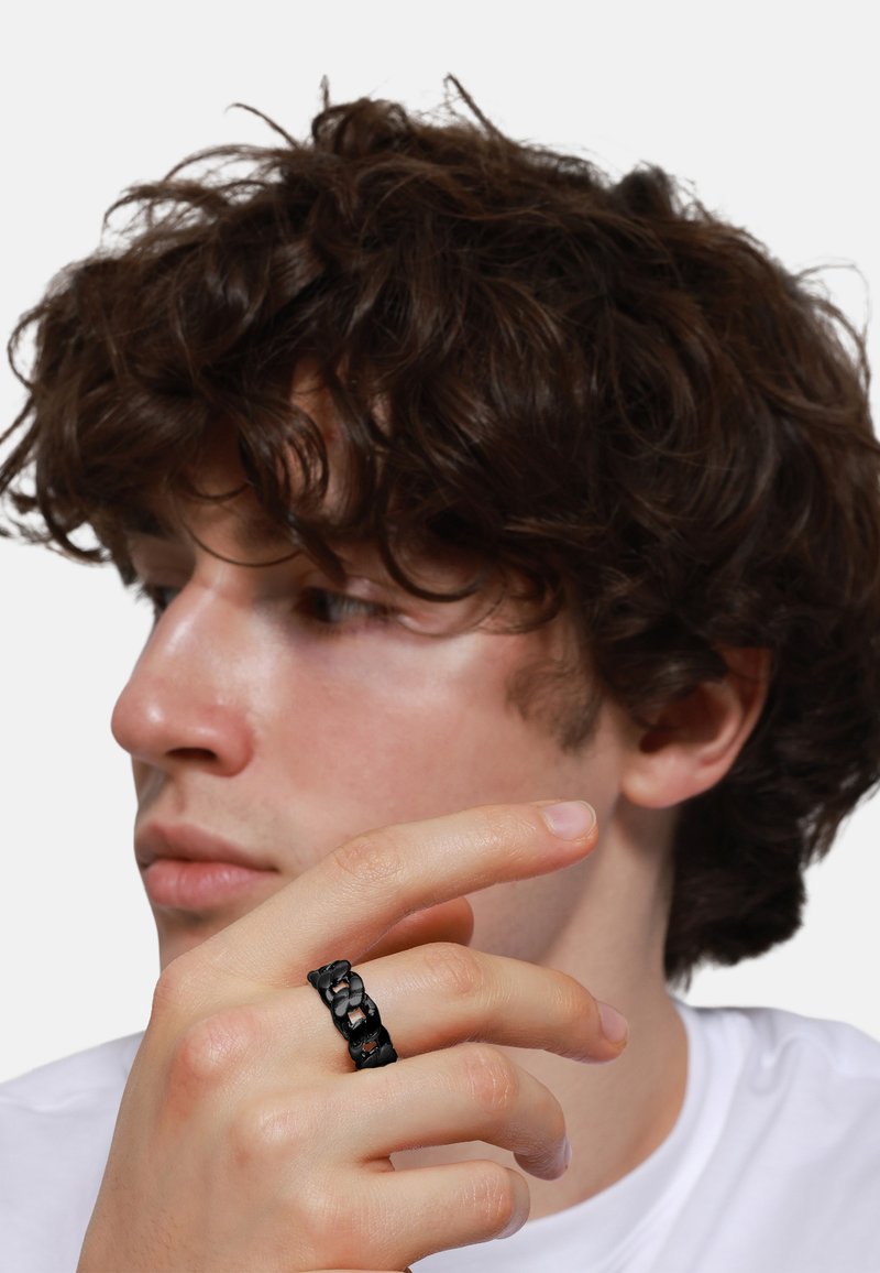 Anello intrecciato nero indossato su una mano con pelle chiara, in contrasto con una maglietta bianca. L'anello presenta un design a catena interconnessa.