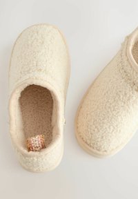 Zapatillas de color crema hechas de un tejido suave y texturizado. Diseño de punta redonda con un interior acolchado y una pequeña etiqueta tejida en la parte posterior.