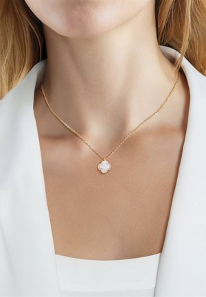 Goldene Kette mit kleinem kleeblattförmigem Perlmuttanhänger, getragen am Hals einer Frau mit weißem Blazer.