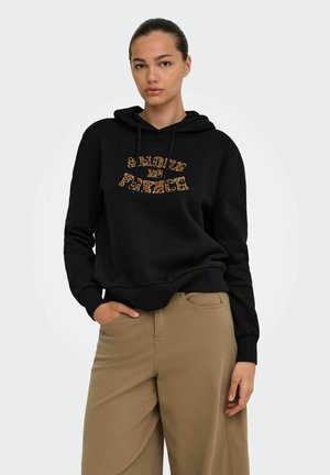 Junge Frau mit zurückgebundenem Haar trägt einen schwarzen Hoodie mit dem Schriftzug „Amour is French“ und beigen Hosen, posiert mit einer Hand in der Tasche.