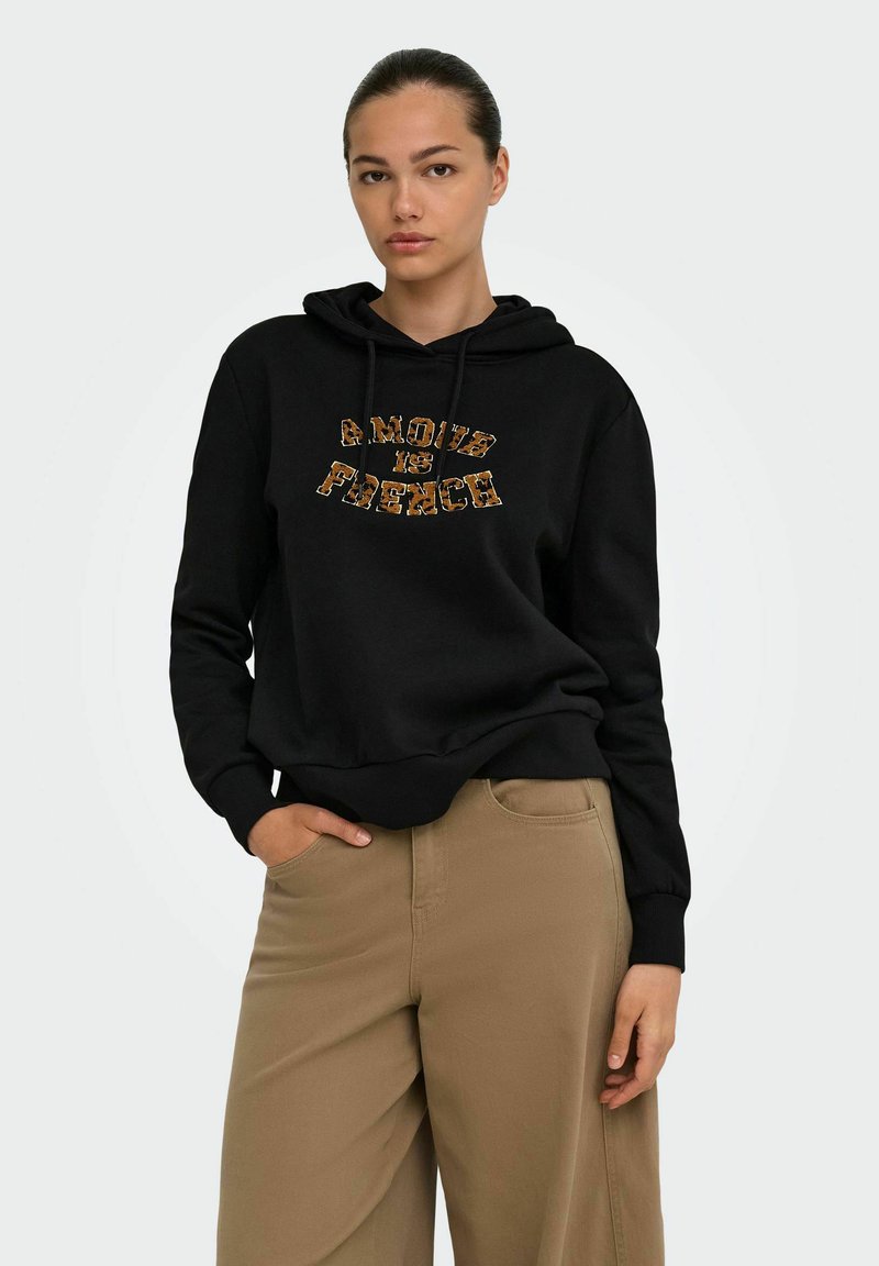 Jeune femme aux cheveux attachés porte un sweat à capuche noir avec l'inscription « Amour is French » et un pantalon beige, posant avec une main dans la poche.