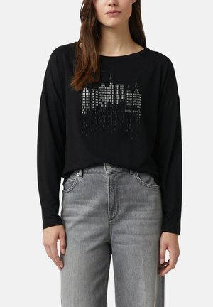 Eine Frau trägt ein schwarzes Langarmshirt mit einem silbernen New Yorker Skyline-Design und hellgrauen, hoch taillierten Jeans.