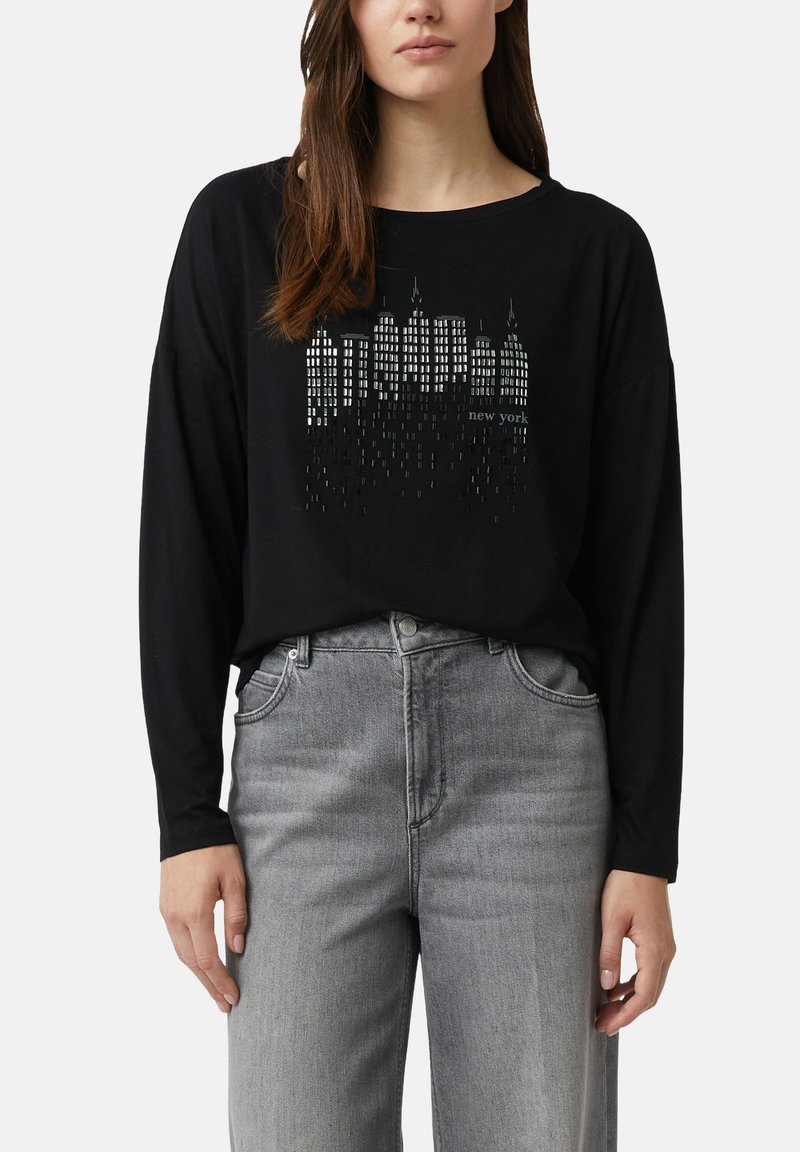 Eine Frau trägt ein schwarzes Langarmshirt mit einem silbernen New Yorker Skyline-Design und hellgrauen, hoch taillierten Jeans.