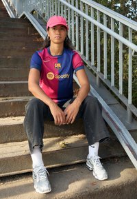 Barcelona FC:n paita sinisenä ja maroonin sävyisenä, yhdistettynä mustiin farkkuihin ja hopeisiin tennareihin. Henkilöllä on pinkki lippis ja valkoiset sukat.