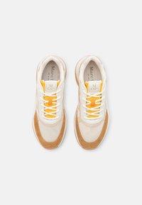 Pár béžových a hnědých tenisek s bílými tkaničkami a žlutými detaily na jazyku, vystavených shora na bílém pozadí.