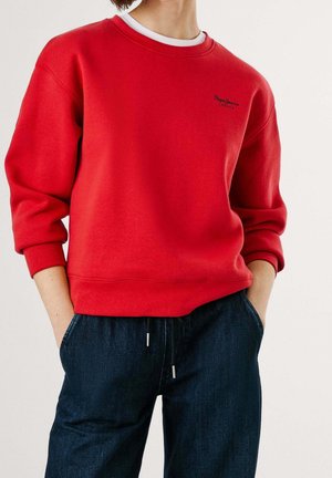 Personne portant un sweat-shirt rouge à manches longues, les mains dans les poches d'un jean bleu foncé.